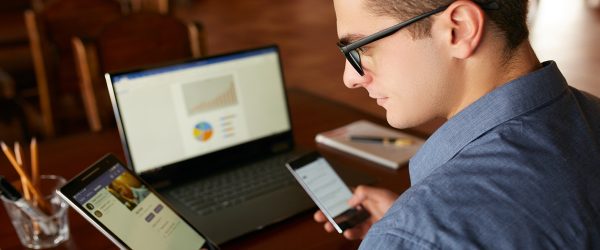 Attractive man in glasses working with multiple electronic devices. Freelancer businessman has laptop and smartphone in hands and laptop on table with charts on screen. Multitasking theme.