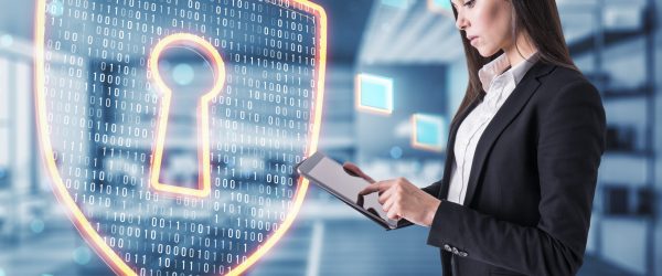 Attractive young european businessman using tablet with glowing shield safety hologram on blurry office interior background. Information and cyber technology concept. Double exposure