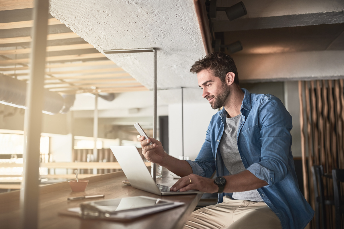 Shot of a handsome young man using a laptop and phone in a coffee shop
