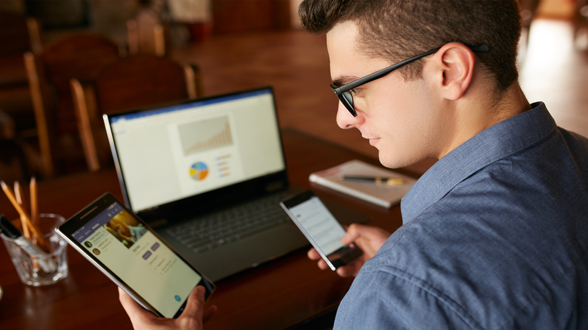 Attractive man in glasses working with multiple electronic devices. Freelancer businessman has laptop and smartphone in hands and laptop on table with charts on screen. Multitasking theme.